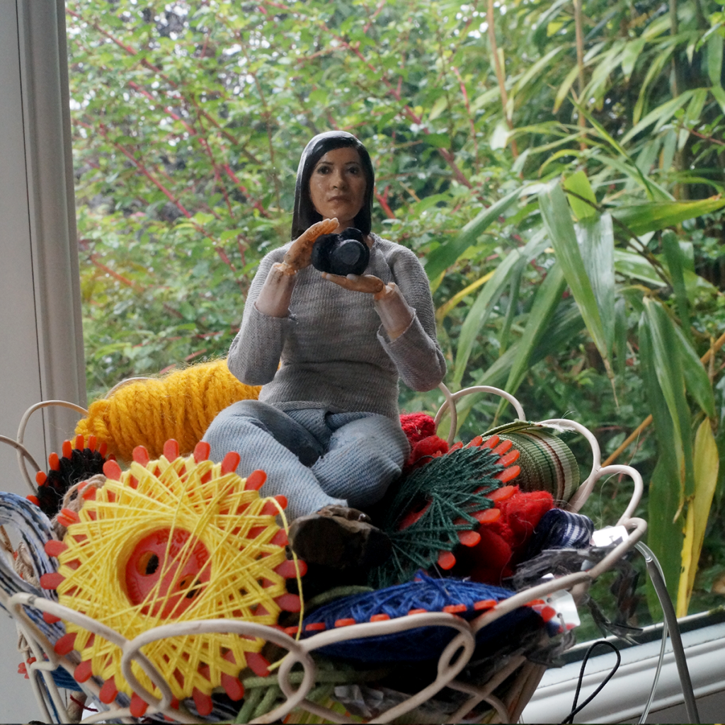 3D printed figure of Lee sitting on thread spools, holding camera.